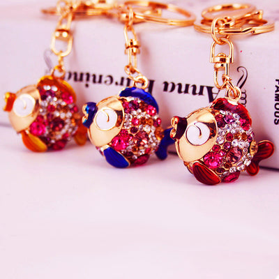 Three rhinestone fish keychains in red, blue, and gold colorways on a pink background.