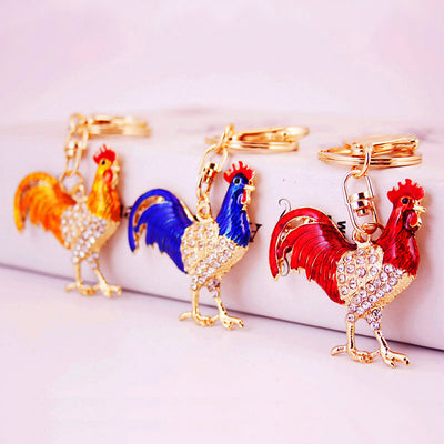 Three gold-tone rooster keychains with gold, blue, and red colored detailing arranged together.