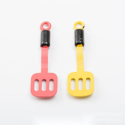 Product view of slotted spatula charm showing a miniature slotted spatula with a black collar and a bright enamel handle