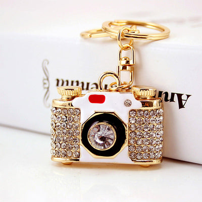 White and gold-tone rhinestone camera keychain displayed beside a book.