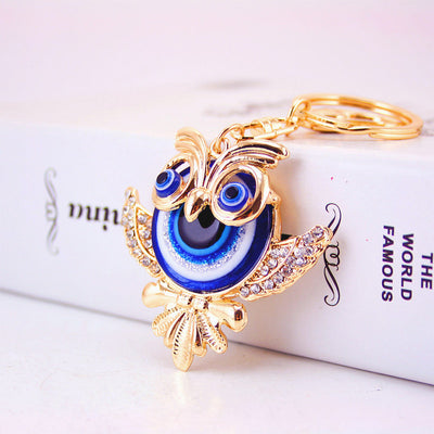 Angled front view of a gold-tone owl keychain with blue eye details and clear stones on the wings.