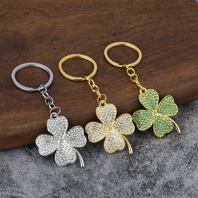 Three clover keychains show silver-tone clear, gold-tone clear, and gold-tone green designs on a wood surface.