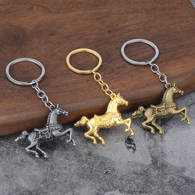 Three saddled horse keychains in silver, gold, and antique bronze tones on a wood and stone surface.