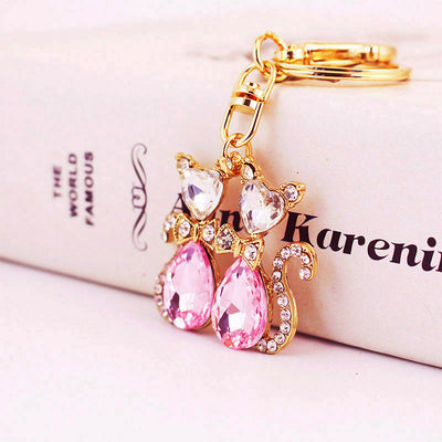 Two gold-tone cat pendants with pink teardrop bodies rest against a white box.