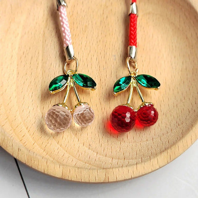 Pink and red faceted crystal cherry phone charms with green leaves on wooden plate