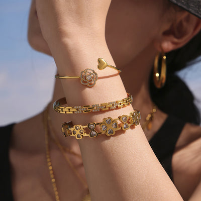 Wrist stacked with gold rose heart open cuff and other gold bracelets outdoors