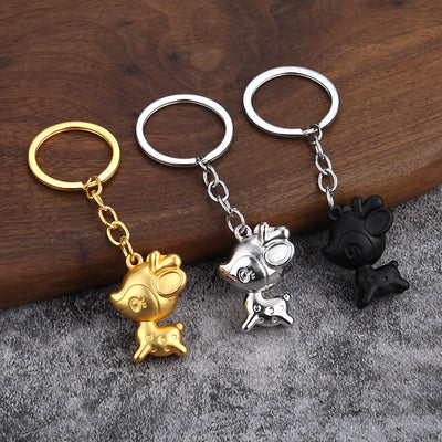 Three cartoon deer keychains in gold, silver, and black on a wood and gray fabric surface.