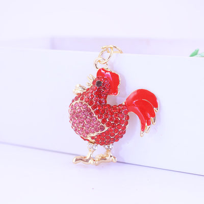 Front view of a red and pink stone-studded rooster charm against a white box.