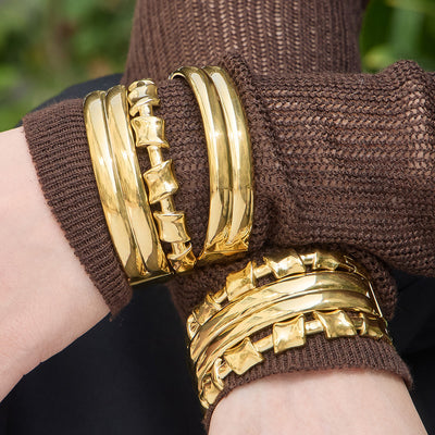 Stacked gold knot-bar and triple-band clasp bangles on brown knit sleeves