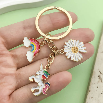 A hand holds a gold-tone keychain with colorful unicorn, rainbow, and daisy charms.