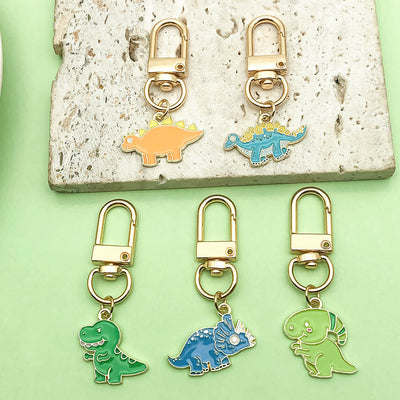 Five colorful dinosaur charms hanging from gold-tone swivel clips on green and stone surfaces.