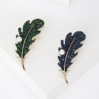 Two colorways of crystal leaf brooch with a slim gold tone vein and white pearl shown side by side.