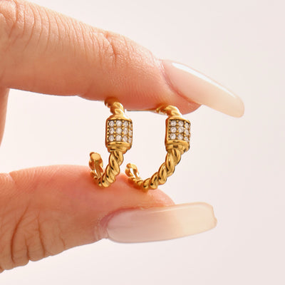 Fingers holding pair of gold twisted C hoop earrings with zircon cubes