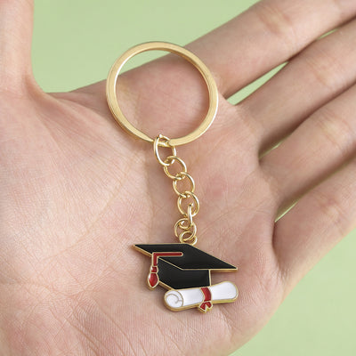 A hand holds a gold-tone key ring with a black graduation cap and white diploma charm.