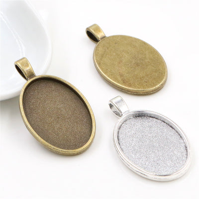 Overview of plain oval bezel pendant trays with broad top bails in silver and antique bronze tones arranged on a light b