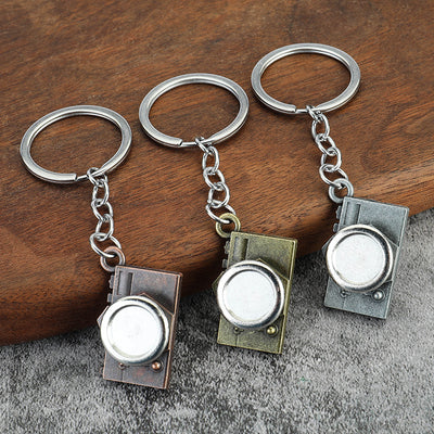 Three camera-shaped keychains in copper, bronze, and silver tones displayed on wood and gray fabric.