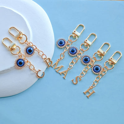 Two gold-tone evil eye charms with letter cutout pendants and swivel clasps on a blue background