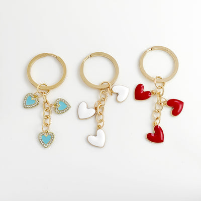 Three gold-tone keychains side by side with turquoise rhinestone, glossy white, and glossy red enamel heart charms.