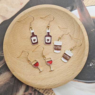 Three pairs of enamel drink earrings wine glasses bottles and coffee cups on wood dish