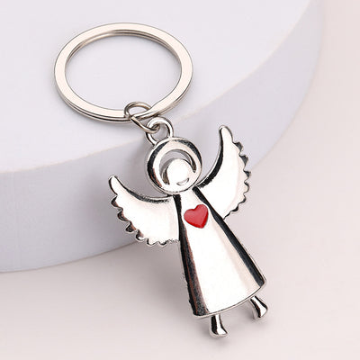 Open-wing silver tone angel keychain with a small red heart on a pale background.