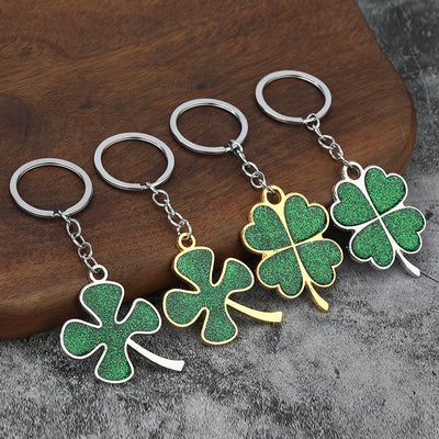 Four green glitter clover keychains in gold-tone and silver-tone frames on a wood and gray surface.
