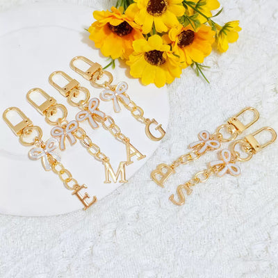 Several gold letter bag charms with pastel enamel bows displayed beside yellow flowers