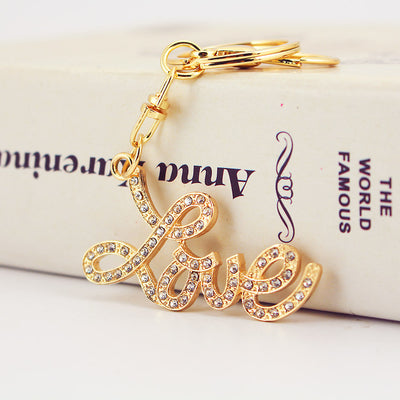 Close-up of a gold-tone Love script charm set with clear rhinestones beside the key ring.