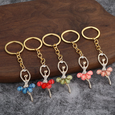 Five ballerina keychains in blue, red, light green, pink, and pale pink are arranged on wood and gray fabric.