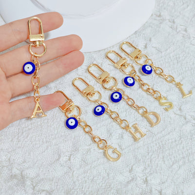 Hand holding two gold letter charm keychains with evil eye bead accents