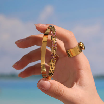 Gold hinged chain-link bangle with pink zircon accents held against beach sky