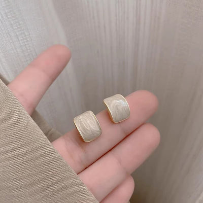 Cream gold-rim square studs held in hand against curtain