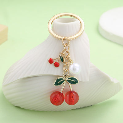 Gold-tone cherry keychain displayed on a white sculpted stand.