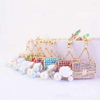 Assorted rhinestone handbag keychains arranged in a row with white flower and pearl-like bead dangles.