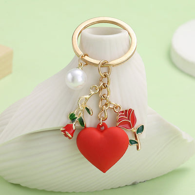 Gold-tone key ring with a red heart, two rose charms and a white bead displayed on a white shell.