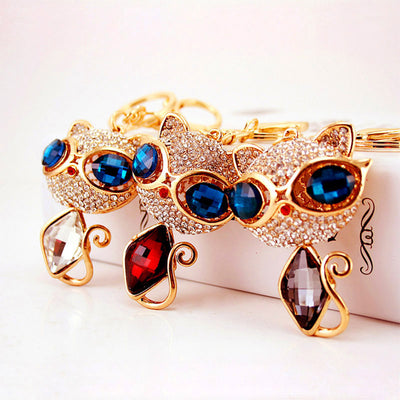 Three gold-tone cat keychains display blue faceted eyes and red, clear, and smoky body stones.