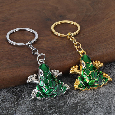 Gold-tone and silver-tone green frog keychains displayed together on a wooden surface.