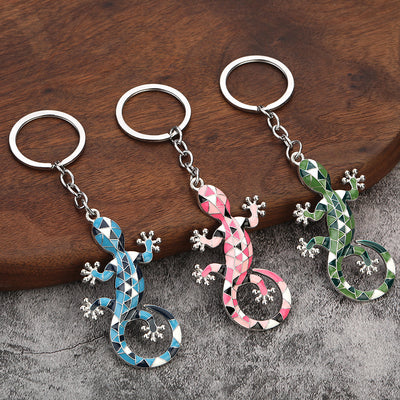 Three gecko keychains in blue, pink, and green on wood and gray fabric.