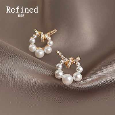 Gold-tone clear-stone bow and pearl-circle stud earrings on pale satin.