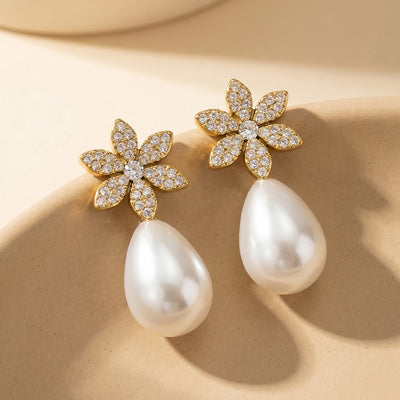 Pair of gold pave flower stud earrings with white teardrop pearl drops on beige surface