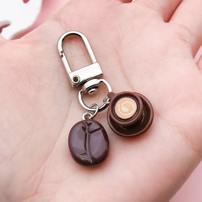 A brown coffee cup and dark coffee bean charm hang from a silver-tone clasp in a hand.