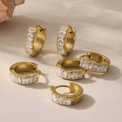 Multiple gold square-zirconia huggie hoop earrings scattered on beige surface