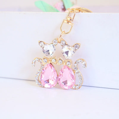 Pink double-cat charm with clear heart-shaped heads, bows, and stone-lined tails.
