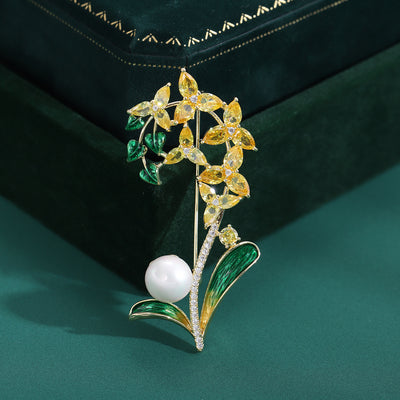 Front view of long orchid-stem brooch with green leaves and pearl-like bead against a white background.