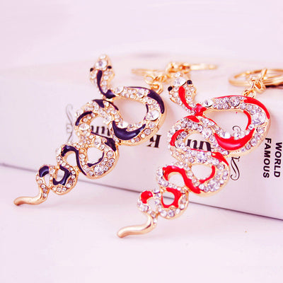 Two gold-tone coiled snake keychains with red and purple accents rest on a pale display box.