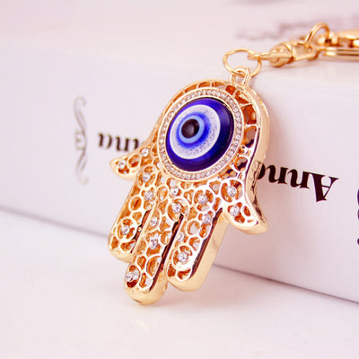 Close-up of a gold-tone Hamsa keychain with a blue eye center and clear stones.