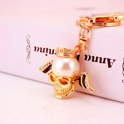 Side view of a gold-tone crowned skull keychain with a pearl-like bead and black rhinestone wings.