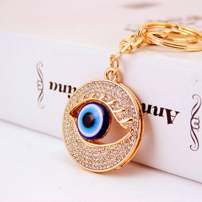 Close-up of a round gold-tone eye pendant with a blue center and rows of clear stones attached to a split key ring.
