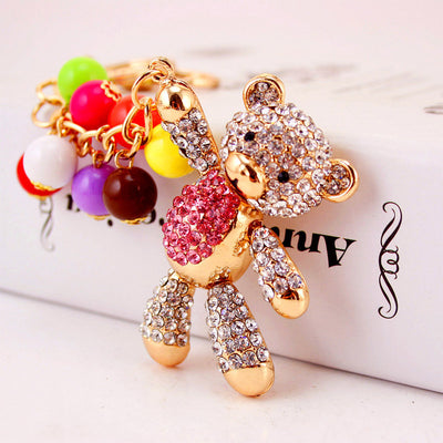 Close-up of a gold-tone teddy bear keychain with a pink rhinestone belly and colorful round beads.