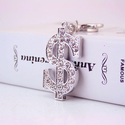 Front close-up of a silver-tone rhinestone dollar sign keychain against a printed book.