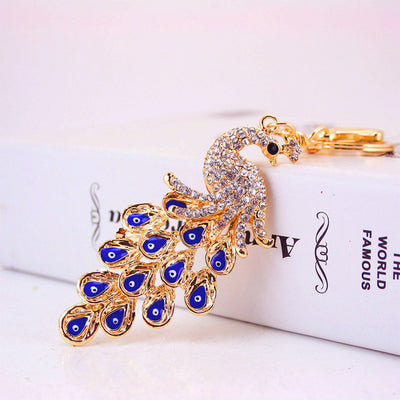 Gold-tone peacock keychain resting against a white book with its blue eye-pattern tail spread out.